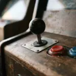 A close-up, low-angle shot of a classic arcade joystick, its well-worn red and blue buttons illuminated by warm
